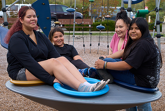 A family of four sit on and stand near a playground spinny saucer, smiling at the camera.