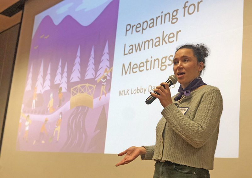 A woman with a microphone gestures and she speaks infront of a projector slide that reads "Preparing for lawmaker meetings"