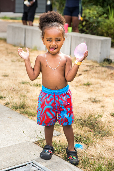 A young child in shorts holds up a water balloon in one hand while sticking out their tongue and making a funny face.