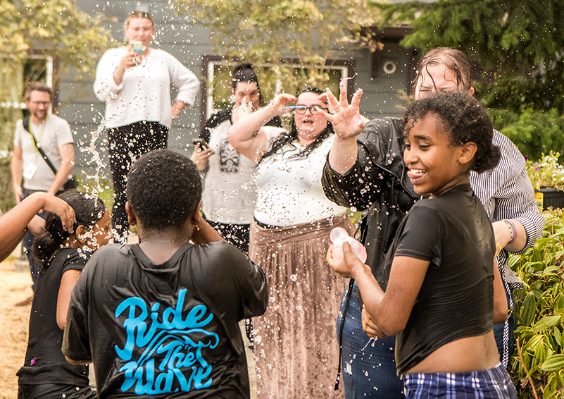 A teenage girl clutches a water balloon amid a crowd of adults and children, all sopping wet, as droplets of water fly through the air