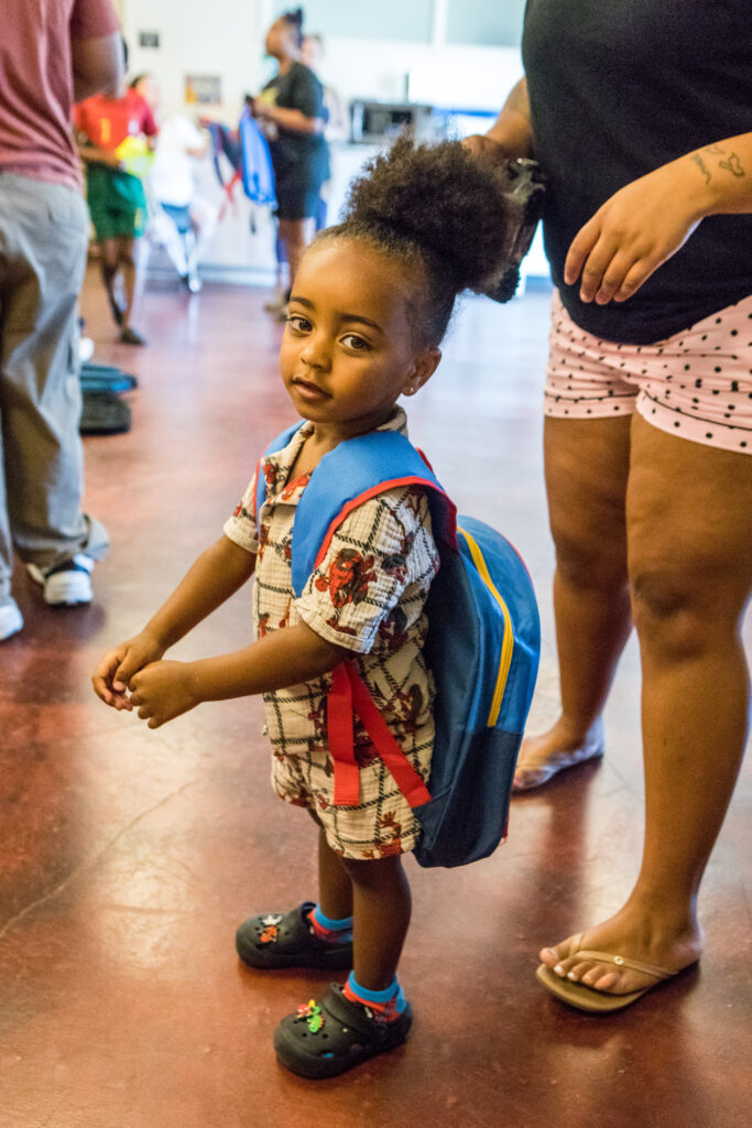 A young boy looks at the camera as he tries on a backpack almost as big as he is.