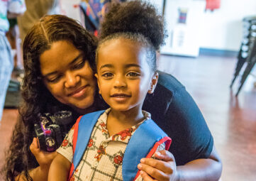 A mom smiles as she crouches down besides her young son wearing a backpack almost as big as he is.