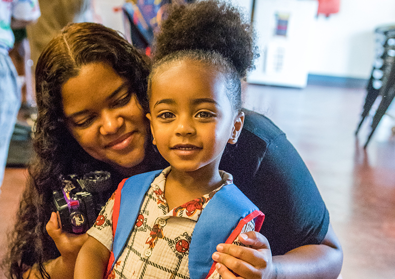 A mom smiles as she crouches down besides her young son wearing a backpack almost as big as he is.