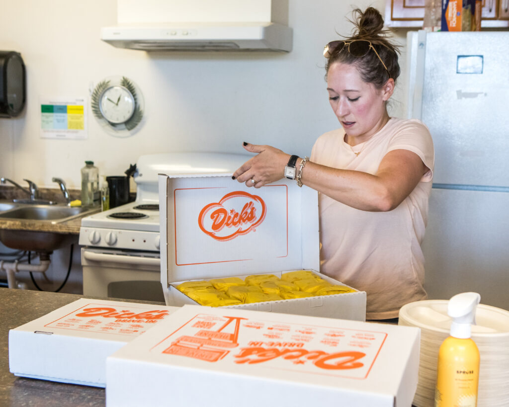 A woman opens a box labeled "Dick's," revealing neat stacks of hamburgers inside.
