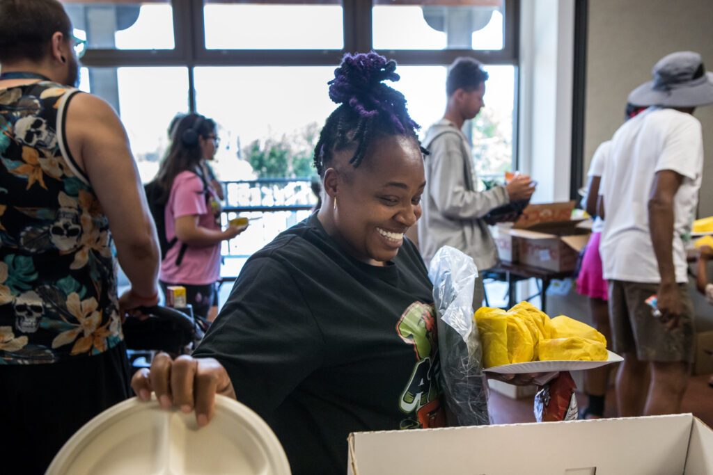 A woman with purple dreds smiles as she holds a plate piled high with hamburgers in one hand and reaches for another plate with the other.