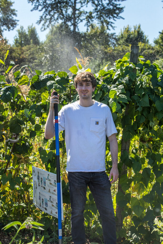 A man in a white T-shirt leans on a shovel while standing in front a vining plant.