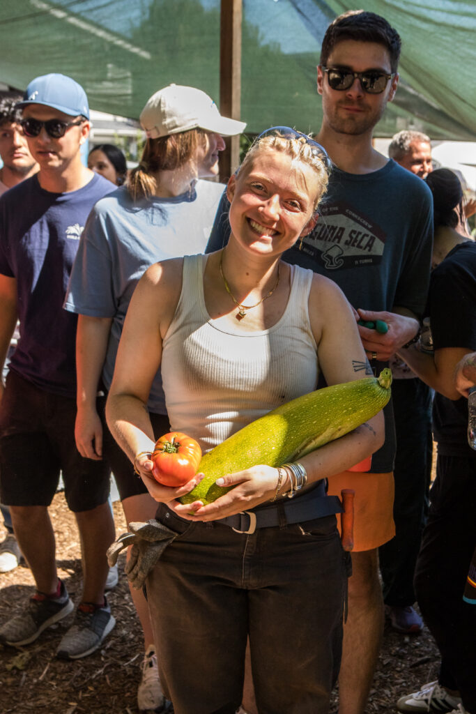 A woman in a white tank top cradles a tomato and an enormous squash.
