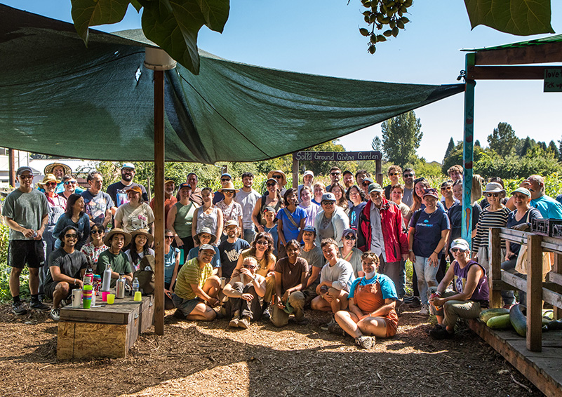 A crowd of people dressed for garden work pose for a group photo under a green cloth canopy.