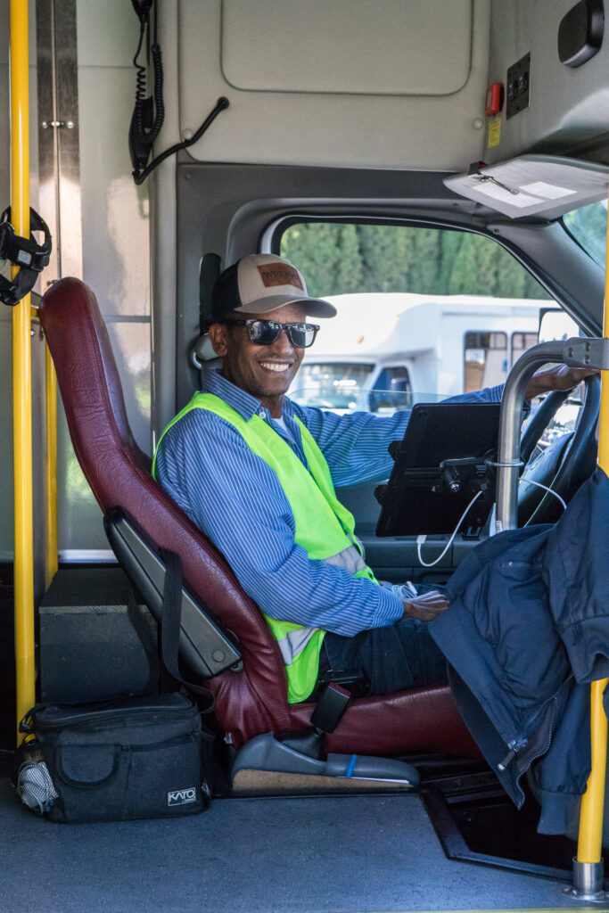 A man in a yellow safety vest, glasses, and a cap sits behind the steering wheel of a bus.