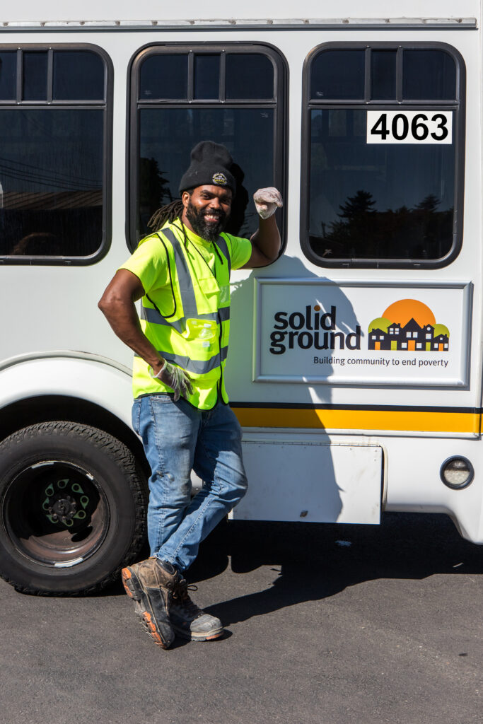 A man in a yellow safety vest leans against a paratransit bus with a sign that reads "Solid Ground."