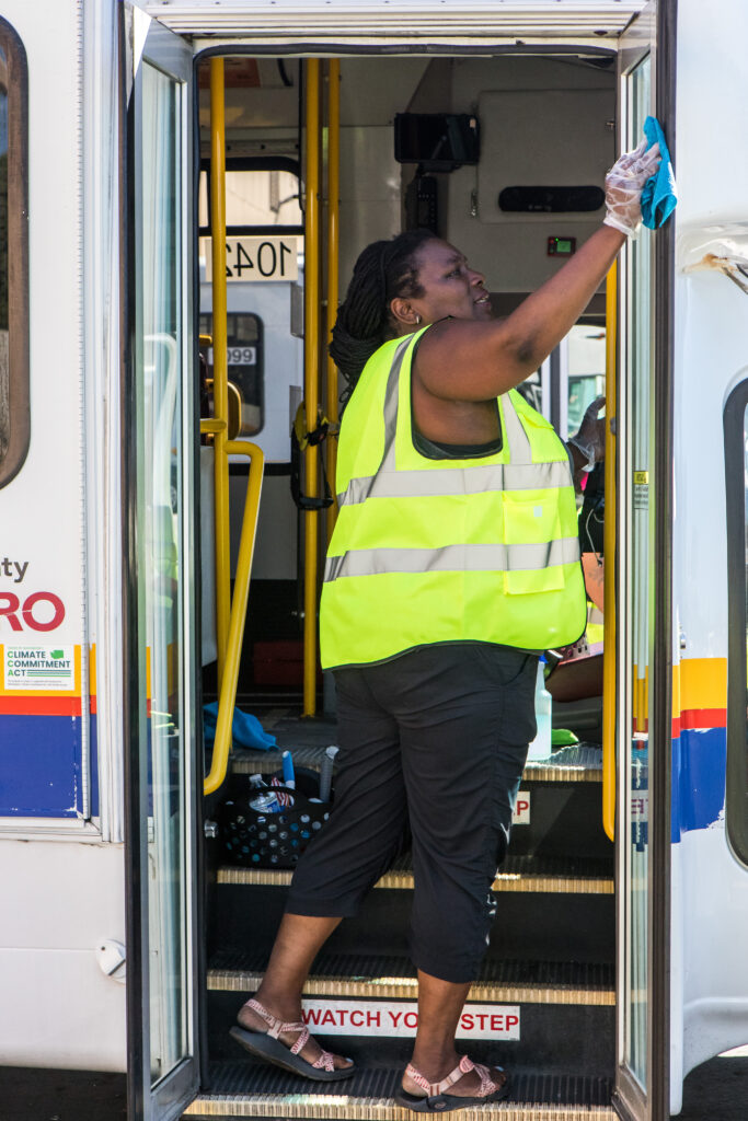 A woman in a yellow safety vest wipes down the door of a paratransit bus.