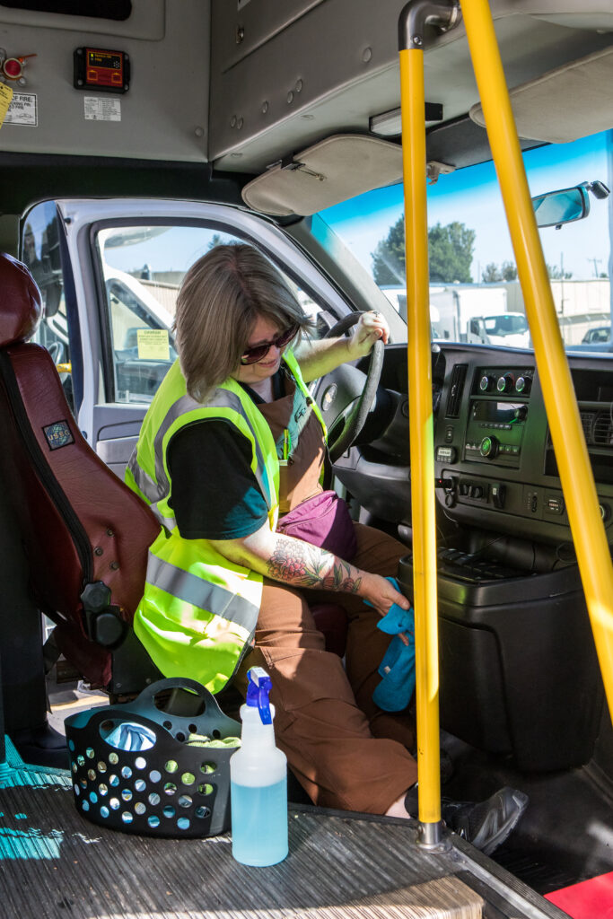 A woman in a yellow safety vest and sunglasses leans down to wipe the consule of a paratransit bus.