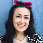 Headshot of a smiling woman with dark brown hair and heart-shaped, red sunglasses on her head, standing in front of a blue painted wall.