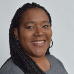Headshot of a smiling woman with long brown braids wearing a grey shirt on a lighter grey background.