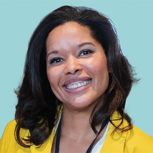 Headshot of a smiling woman with brown hair wearing a bright yellow blazer on a light blue background.
