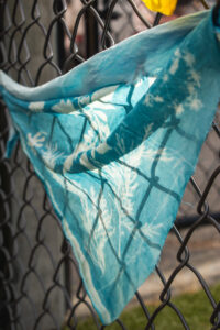 A triangle of blue cloth imprinted with white shapes of leaves and flowers hangs from a chainlink fence.