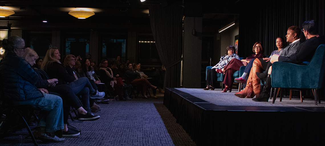 Five women of color sit on a stage in front of a large audience.