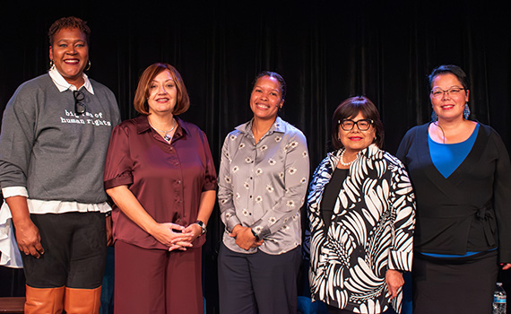 Five women of color pose smiling on a stage in front of a dark curtain.