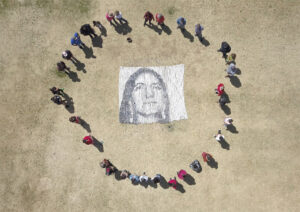 An arial shot of people standing in a circle in a field around a mosaic of a face made up large clay beads of various shades of gray.