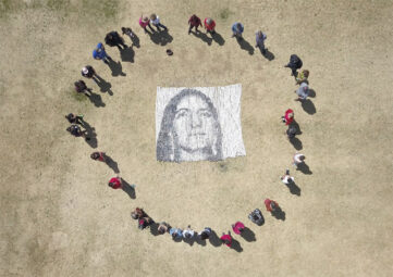 An arial shot of people standing in a circle in a field around a mosaic of a face made up large clay beads of various shades of gray.