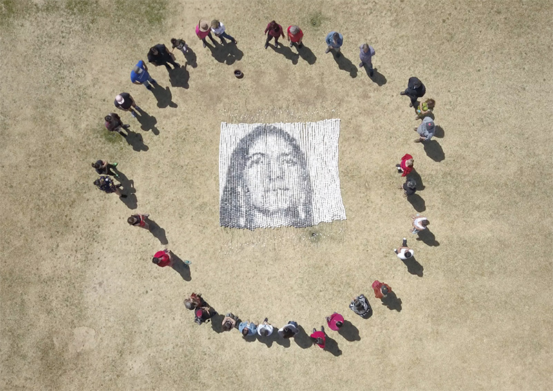 An arial shot of people standing in a circle in a field around a mosaic of a face made up large clay beads of various shades of gray.