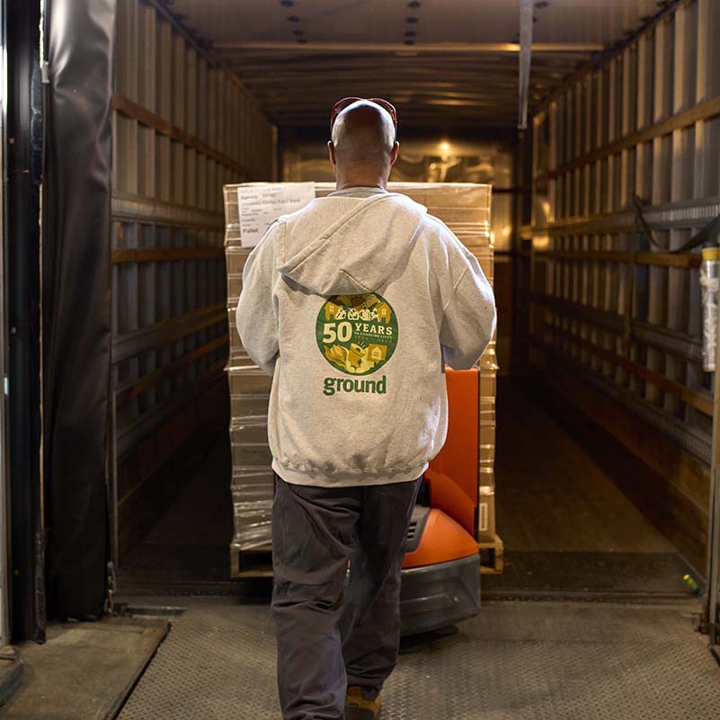 A man wearing a gray sweater with a Solid Ground logo on the back is seen from behind wheeling a pallet of food into the back of a truck.