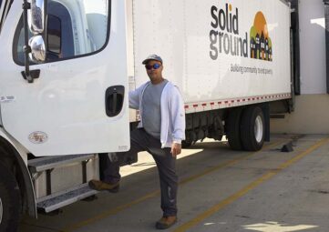 A man wearing a hat and a sweater stands behind the open cap of a white box truck with the Solid Ground logo painted on the side.