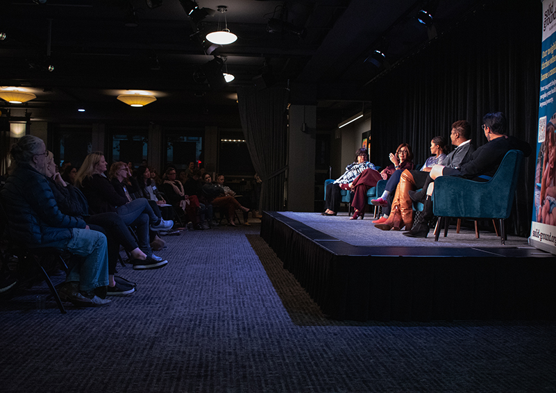 Five women sit on chairs on a stage acros from a packed audience in a darkened room.