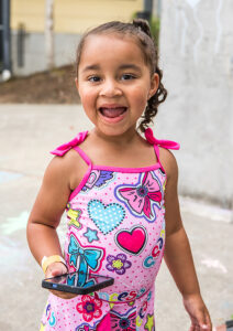 A little girl in a pink bathing suit grins at the camera. She's holding her mom's cellphone.