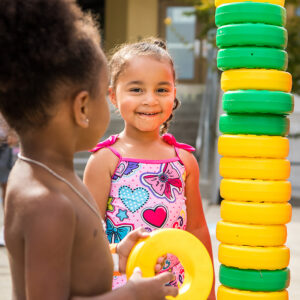 Two young kids in bathing suits stand next to a tower of green and yellow plastic rings.