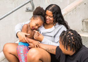 Two women tickle a little boy as they all sit on a set of concrete steps.