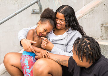 Two women tickle a little boy as they all sit on a set of concrete steps.