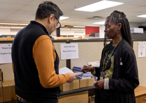 A man with dark hair and glasses looks at a booklet as he chats with a woman with long dark braids.