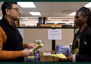 A man with dark hair and glasses looks at a booklet as he chats with a woman with long dark braids.