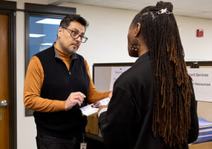 A man with dark hair and glasses looks at a booklet as he chats with a woman with long dark braids.