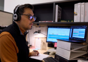 A man with dark hair, wearing glasses and a headset, sits in front of multiple computer monitor as he presents a webinar.