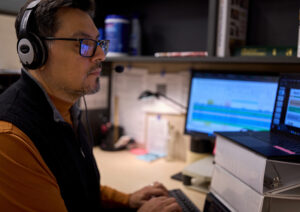 A man with dark hair, wearing glasses and a headset, sits in front of multiple computer monitor as he presents a webinar.