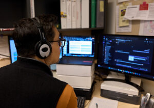A man with dark hair, wearing glasses and a headset, sits in front of multiple computer monitor as he presents a webinar.