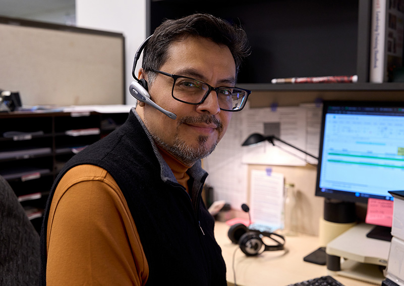 A man with dark hair, wearing glasses and a headset, sits in front of a computer monitor and smiles at the camera.