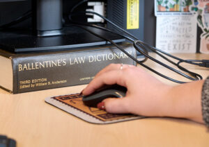 A woman's hand rests on a computer mouse in front of a computer monitor raised up on a thick book titled BALLENTINE'S LAW ENCYCLOPEDIA.