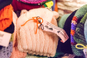 A pink knitted hat with a Solid Ground/AmeriCorps Seniors tag, surrounded by other colorful knitted hats.