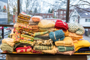 Knitted blankets and hats stacked on a table in front of a large window, with buildings visible outside.