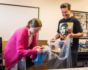 A young woman with blonde hair, glasses, and a bright pink sweater, drops knitted hats into a plastic bag held by a man with brown hair and black T-shirt.