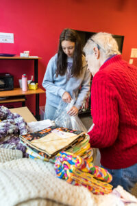 Two women, one young with long brown hair and one older with short white hair, pack knitted items into large plastic bags.