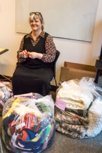 A woman sits on a chair surrounded by several large plastic bags filled with knitted items.