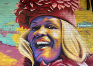 A colorful mural of a smiling woman with blonde hair and dark pink featured hat. Also in the mural is a street sign that reads "DeCharlene".
