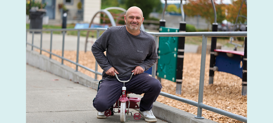 A balding man with grey facial hair sits on a tiny red tricycle in front of playground equipment and smiles.