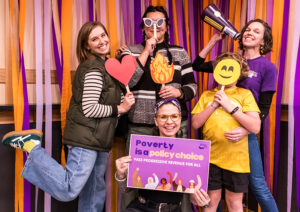 A group of people making goofy poses infront of orange and purple streamers while holding props -- including a heart and smiley face -- along with a sign th at reads "Poverty is a policy choice."