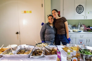 Two women embrace behind a table piled high with food.