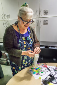 A smiling woman in a colorful apron arraning craft supplies on a table.